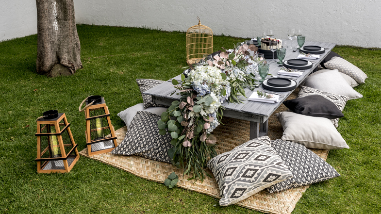 Montaje de picnic elegante en el jardín con mesa baja decorada con arreglos florales, cojines decorativos y linternas de madera. Ideal para renta y venta en eventos especiales